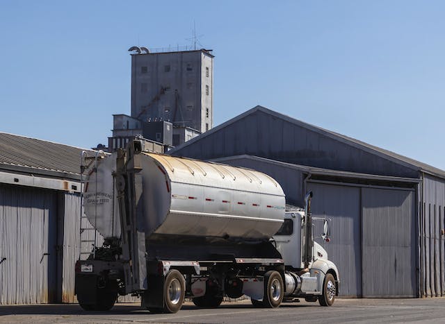 A white tanker truck parked beside industrial buildings with a grain elevator in the background under a clear blue sky.